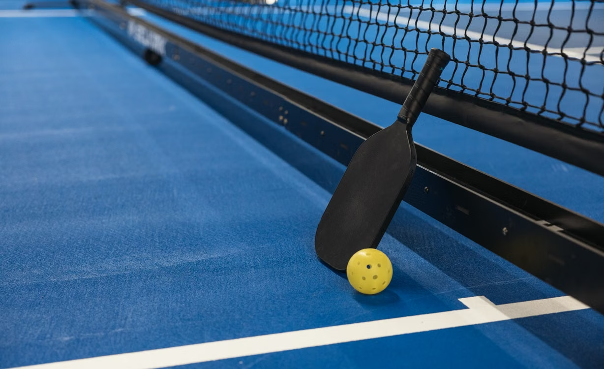 Pickleball paddle and yellow ball resting beside a net on a blue court